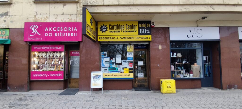 Zdjęcie przedstawia trzy sklepy. Po lewej stronie znajduje się różowy szyld z napisem "AKCESORIA DO BIŻUTERII" oraz "ROYAL STONE". Po środku znajduje się sklep z żółtym szyldem Cartridge Center. Sprzedaje on tusze do drukarek w Warszawie, tonery Warszawa. Po prawej stronie sklep z używaną odzieżą.